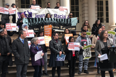 A group of protesters stand outside with signs from "Make the Road", "JFREJ", "NYC Against Hate", etc.