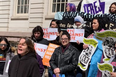 A group of protesters stand outside with signs from "Make the Road", "JFREJ", "NYC Against Hate", etc.