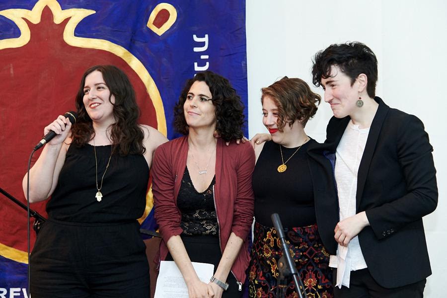 Four women stand in front of a banner while one speaks into a microphone