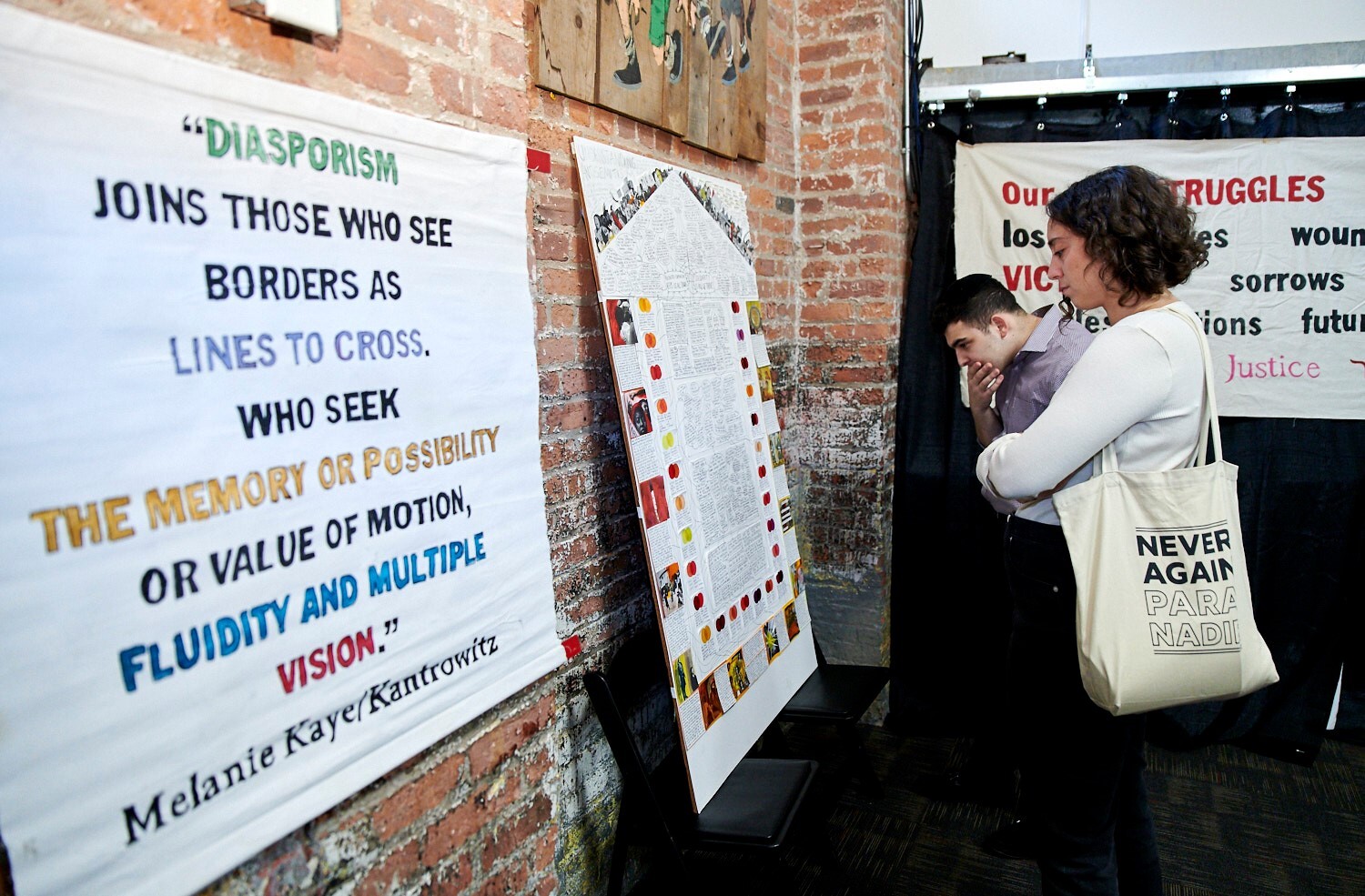 Image of two people looking at a banner and a poster. The banner reads "Diasporism joins those who see borders as lines to cross. Who seek the memory or possibility or value of motion, fluidity and multiple vision. - Melanie Kaye/Kantrowitz"